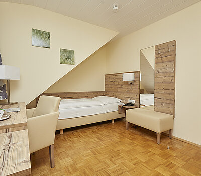 Das Foto zeigt ein Einzelzimmer mit einem Holzboden. Links steht ein kleiner Holztisch mit einem beigen Sessel. Auf dem Tisch steht ein Tablett mit einer Kaffeetasse. Das Bett ist in eine Nische unter einer abgeschrägten Decke eingebaut und hat eine beige Lehne. Rechts neben dem Bett befindet sich eine kleine Bank und ein großer Spiegel an einer mit Holz verkleideten Wand. An der Wand hängen zwei kleine Bilder. Rechts im Bild ist eine Holztür zu sehen.