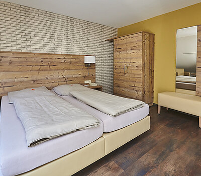 Das Foto zeigt ein Doppelzimmer. Das Bett hat zwei separate Matratzen. Rechts im Raum steht ein Schrank mit einer integrierten Garderobe, einer Kofferablage und einem Spiegel. Der Boden ist aus dunkelbraunem Holz.