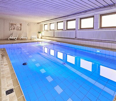Helles Hallenschwimmbad mit einem großen, rechteckigen Pool, umgeben von Fenstern und einer hellen holzdecke Decke mit Beleuchtung.