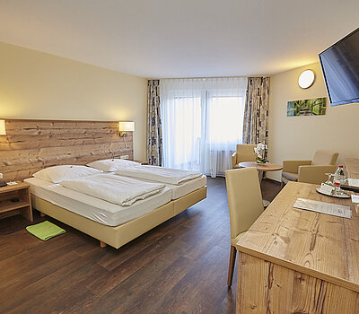 Das Standardzimmer ist ausgestattet mit einem Holzbett mit beiger Lehne. Rechts davon steht ein Holztisch in Beige, darüber hängt ein Flachbildfernseher. Das Zimmer hat Zugang zur Terrasse durch große Türen.