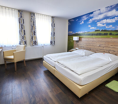 Das Foto zeigt ein Doppelzimmer mit einem Doppelbett. Das Bett hat eine Lehne aus Holz. An der Wand hinter dem Bett befindet sich eine Tapete mit einer Ansicht auf eine mittelfränkische Wiese.
