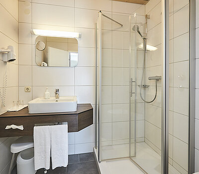 Das Foto zeigt ein Badezimmer mit einem Waschbecken und einer bodengleichen Dusche. Es ist ausgestattet mit einem Föhn, einem Spiegel, Handtüchern und großen weißen Kacheln.