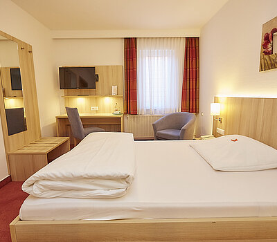 Das Foto zeigt ein Einzelzimmer mit einem Holzboden. Links steht ein kleiner Holztisch mit einem beigen Sessel. Auf dem Tisch steht ein Tablett mit einer Kaffeetasse. Das Bett ist in eine Nische unter einer abgeschrägten Decke eingebaut und hat eine beige Lehne. Rechts neben dem Bett befindet sich eine kleine Bank und ein großer Spiegel an einer mit Holz verkleideten Wand. An der Wand hängen zwei kleine Bilder. Rechts im Bild ist eine Holztür zu sehen.