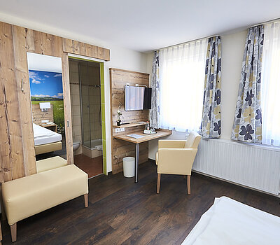 Das Foto zeigt ein Zimmer mit einer Garderobe in Holzfassade. Die Garderobe hat eine Kofferablage und einen großen Spiegel und befindet sich gegenüber dem Bett. Rechts im Raum sind zwei Fenster mit langen Heizkörpern darunter. Davor steht ein kleiner Schreibtisch mit einem beigen Sessel.